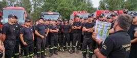 Kilkudziesiecioosobowa grupa strażaków podczas zbiórki. W tle widoczne ustawione samochody pożarnicze. Stojący przed wszystkimi na pierwszym planie dowódca trzyma w ręku kolorowy harmonogram