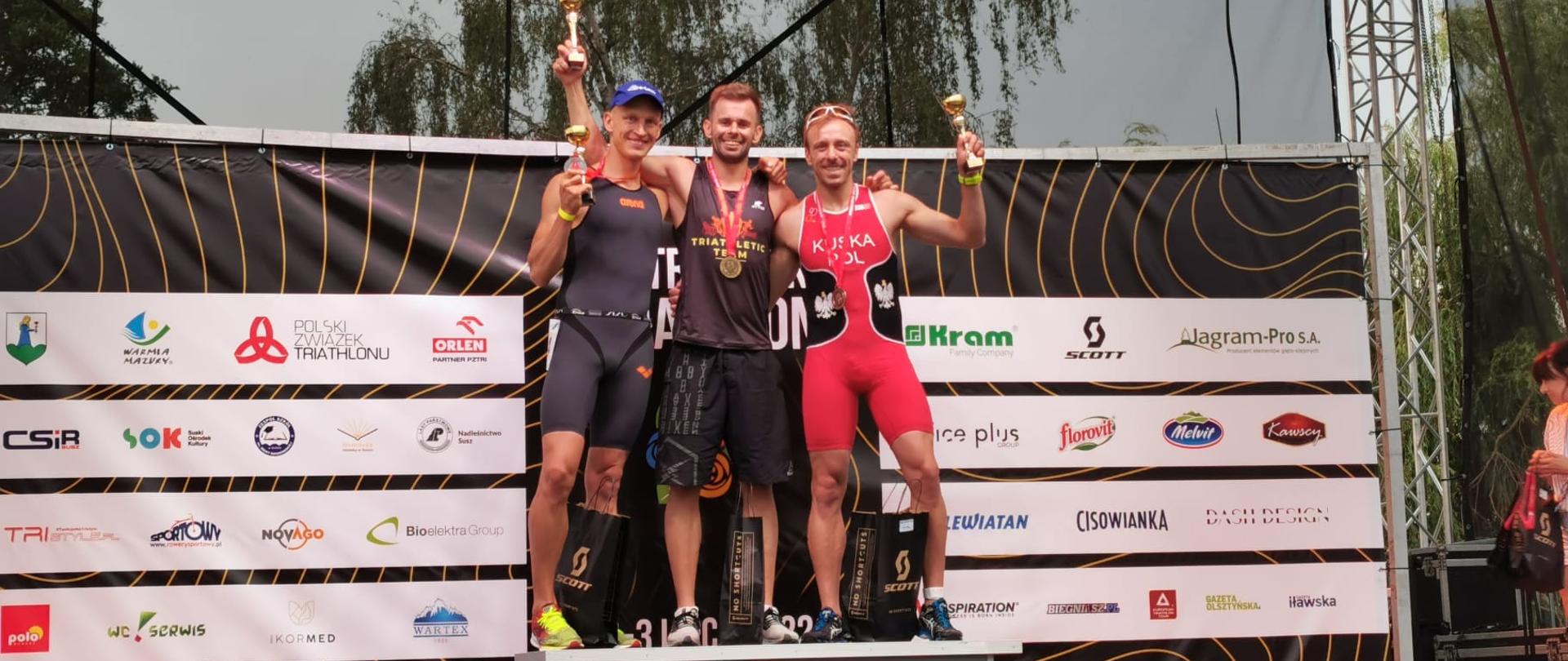 trzech mężczyzn w obcisłych strojach stojących na podium w rękach trzymają puchary