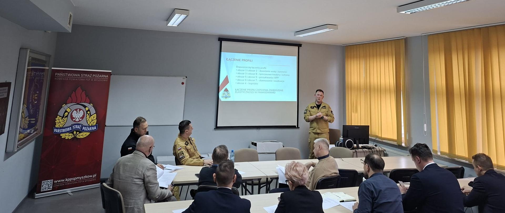 Sala konferencyjna w Komendzie Powiatowej PSP w Myszkowie. Przy stołach siedzą strażacy oraz przedstawiciele samorządów, przeglądający dokumenty. Z przodu sali funkcjonariusz w umundurowaniu PSP prowadzi prezentację wyświetlaną na ekranie. Po lewej stronie widoczny jest roll-up z logo Państwowej Straży Pożarnej, a po prawej duże okna z żółtymi roletami.