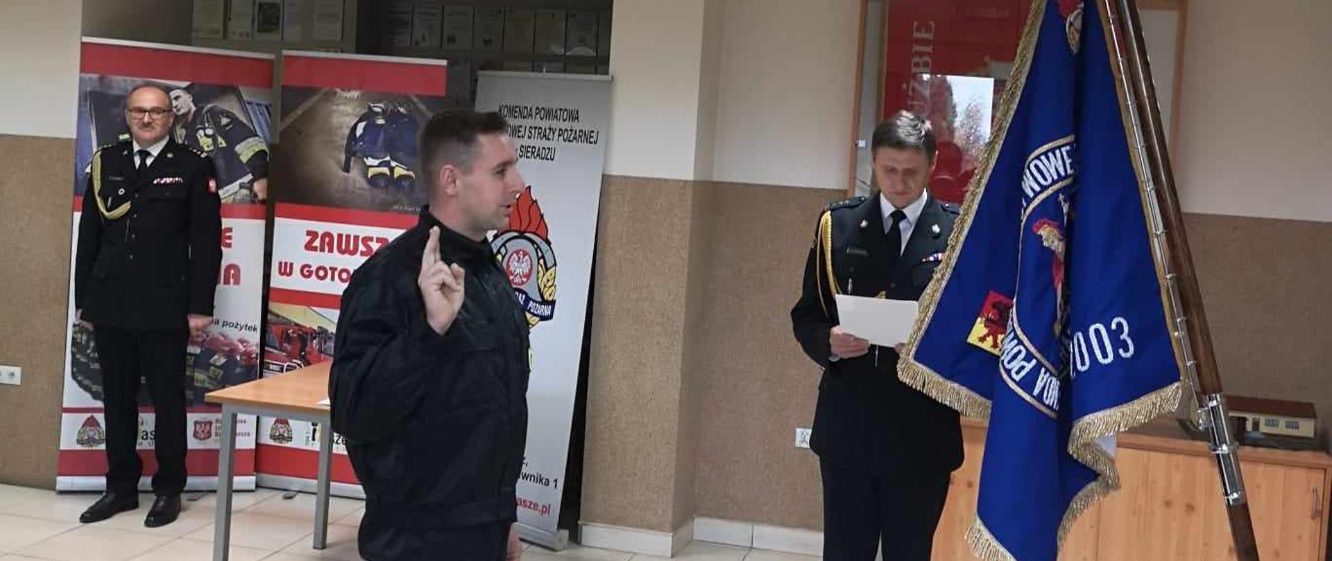 Na zdjęciu widzimy strażaka który składa słowa ślubowania. Po prawej stronie widzimy Sztandar Komendy Powiatowej Państwowej Straży Pożarnej w Sieradzu. Na drugim planie znajdują się Komendanci Powiatowi. 