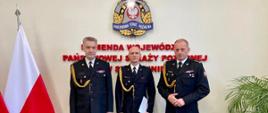 Trzech mężczyzn w mundurach, na tle ściany z logiem i napisem "Komenda Wojewódzka Państwowej Straży Pożarnej.
Na zdjęciu widoczne flagi Polski oraz roślina.