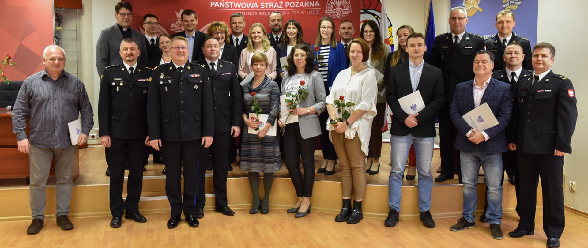 pracownicy cywilni, funkcjonariusze oraz komendanci komendy wojewódzkiej PSP w Łodzi pozują do zdjęcia w sali konferencyjnej, za nimi a tle ścianka reklamowa komendy