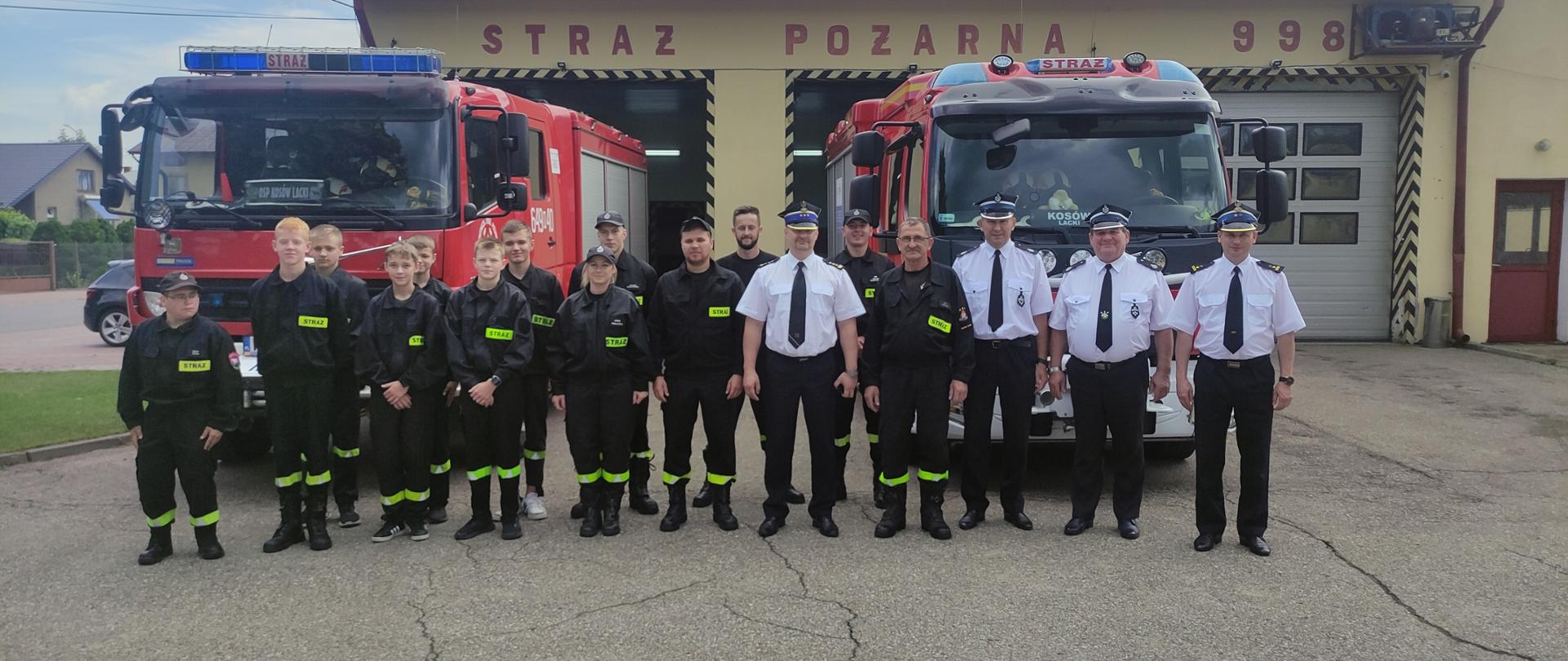 Wizytacja Zastępcy Mazowieckiego Komendanta Wojewódzkiego PSP w powiecie sokołowskim - na zdjęciu strażacy na zbiórce na placu przed strażnicą przed czerwonymi samochodami strażackimi w tle remiza.