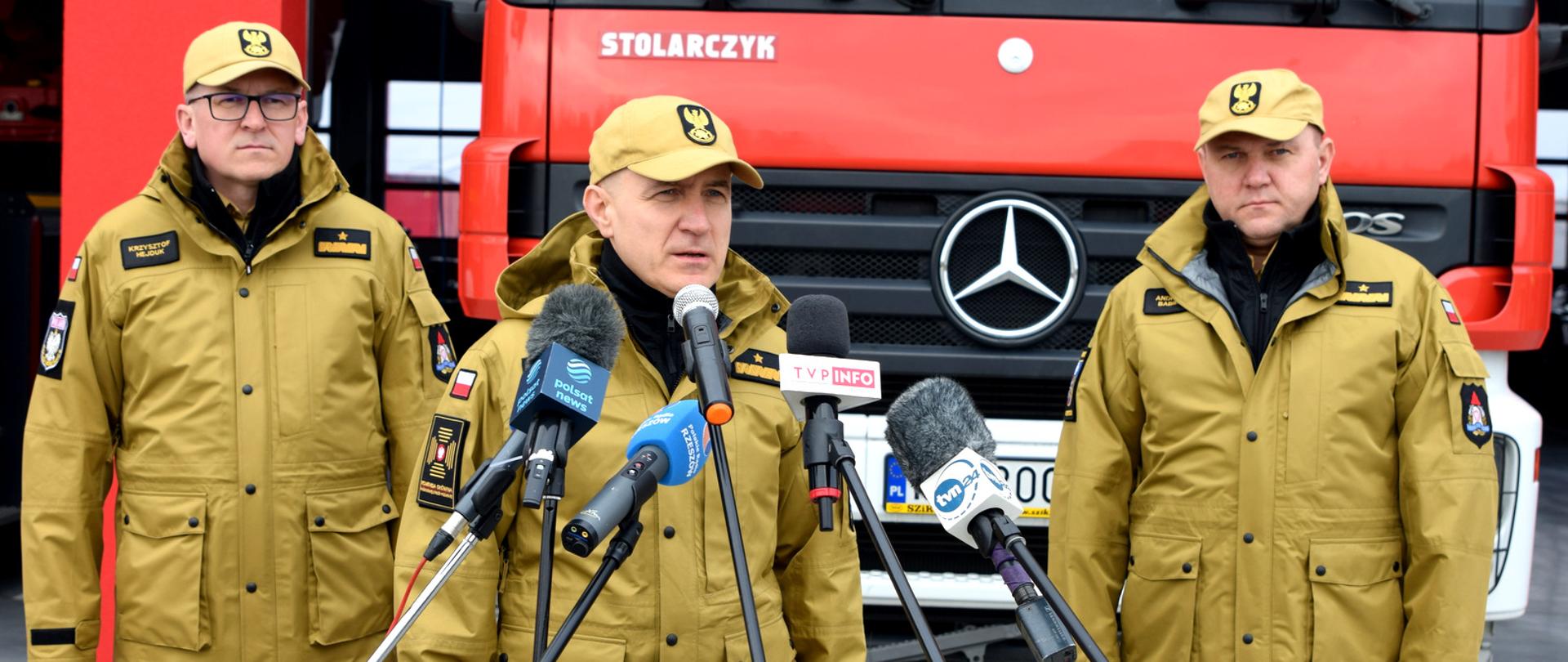Zdjęcie wykonano na zewnątrz. Na zdjęciu trzech oficerów Państwowej Straży Pożarnej w stopniach Generałów. Oficerowie uczestniczą w konferencji prasowej. Mężczyźni ustawieni są w szeregu. Jeden z nich przemawia do zestawu mikrofonów. Za strażakami widać przód samochodu gaśniczego. 