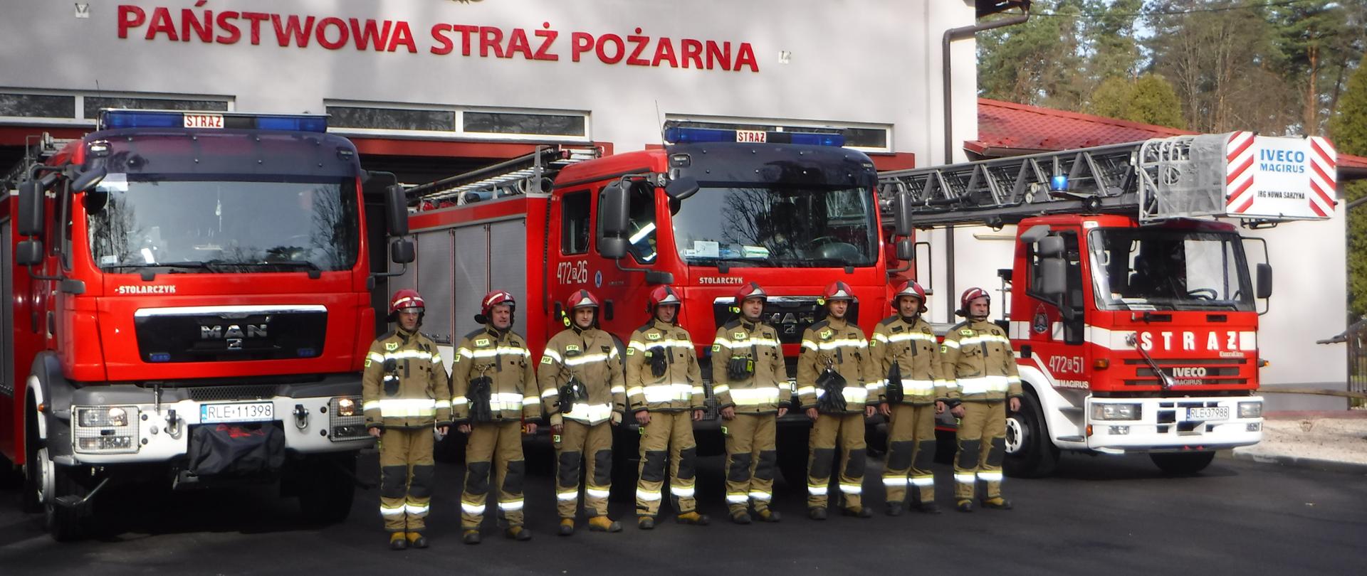 Na zdjęciu widzimy strażaków, którzy stoją na tle pojazdów pożarniczych w tle widać budynek jednostki ratowniczo gaśniczej. 