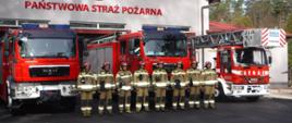 Na zdjęciu widzimy strażaków, którzy stoją na tle pojazdów pożarniczych w tle widać budynek jednostki ratowniczo gaśniczej. 