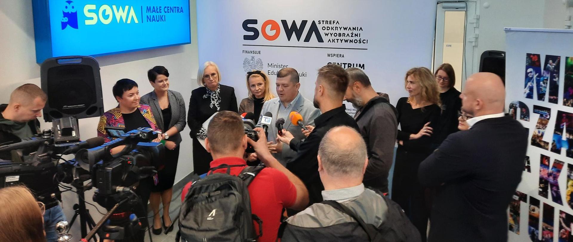 Pod ścianą z napisem SOWA stoi duża grupa ludzi, mężczyzna w szarej marynarce mówi do kilku mikrofonów trzymanych przed dziennikarzy.