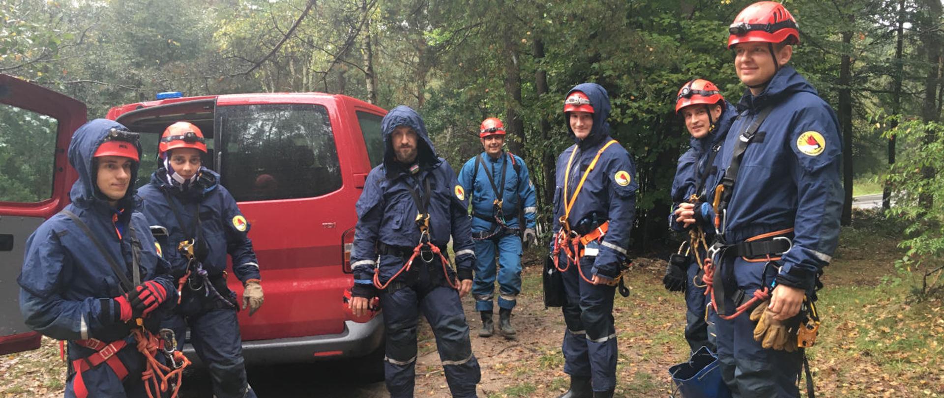 Grupa siedmiu mężczyzn-członków sekcji wysokościowej stojąca na leśnej drodze, w głębi widoczny tył samochodu typu van minibus