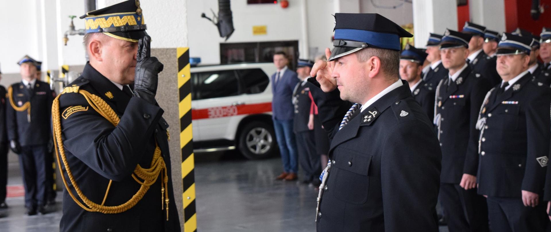 Zdjęcie zrobione wewnątrz garażu jednostki ratowniczo-gaśniczej. Po lewej stronie generał PSP w mundurze galowym salutuje, po prawej stronie strażak ochotni w mundurze galowym salutuje. W tle zebrani na zbiórce strażacy PSP i OSP.