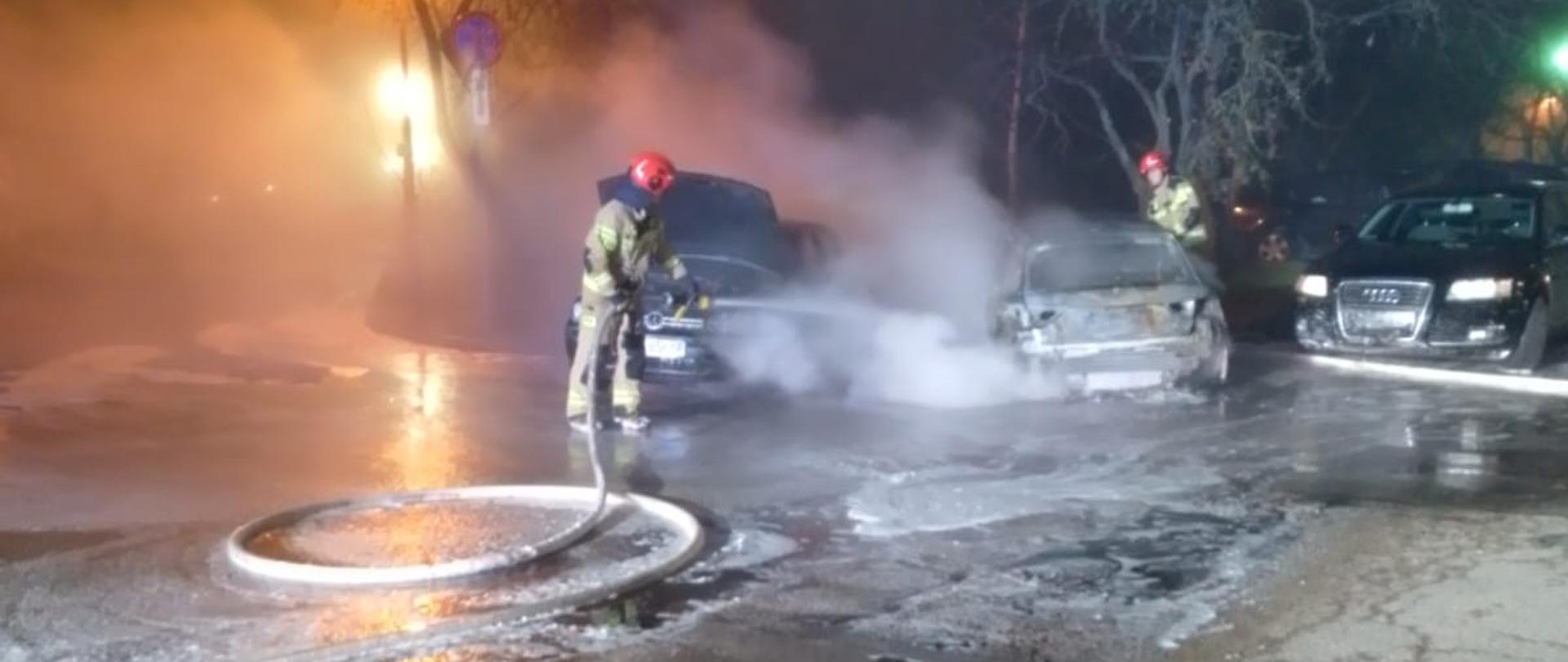 Zdjęcie przedstawia ratownika, który strumieniem piany gasi palące się samochody. 