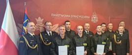 Pamiątkowe zdjęcie grupowe w sali Państwowej Straży Pożarnej w Szczecinie. Oficerowie w galowych mundurach oraz nowo przyjęci strażacy w czarnych koszulach z napisem STRAŻ pozują razem, trzymając w rękach białe teczki z dokumentami. Po lewej stronie widoczna jest flaga Polski.