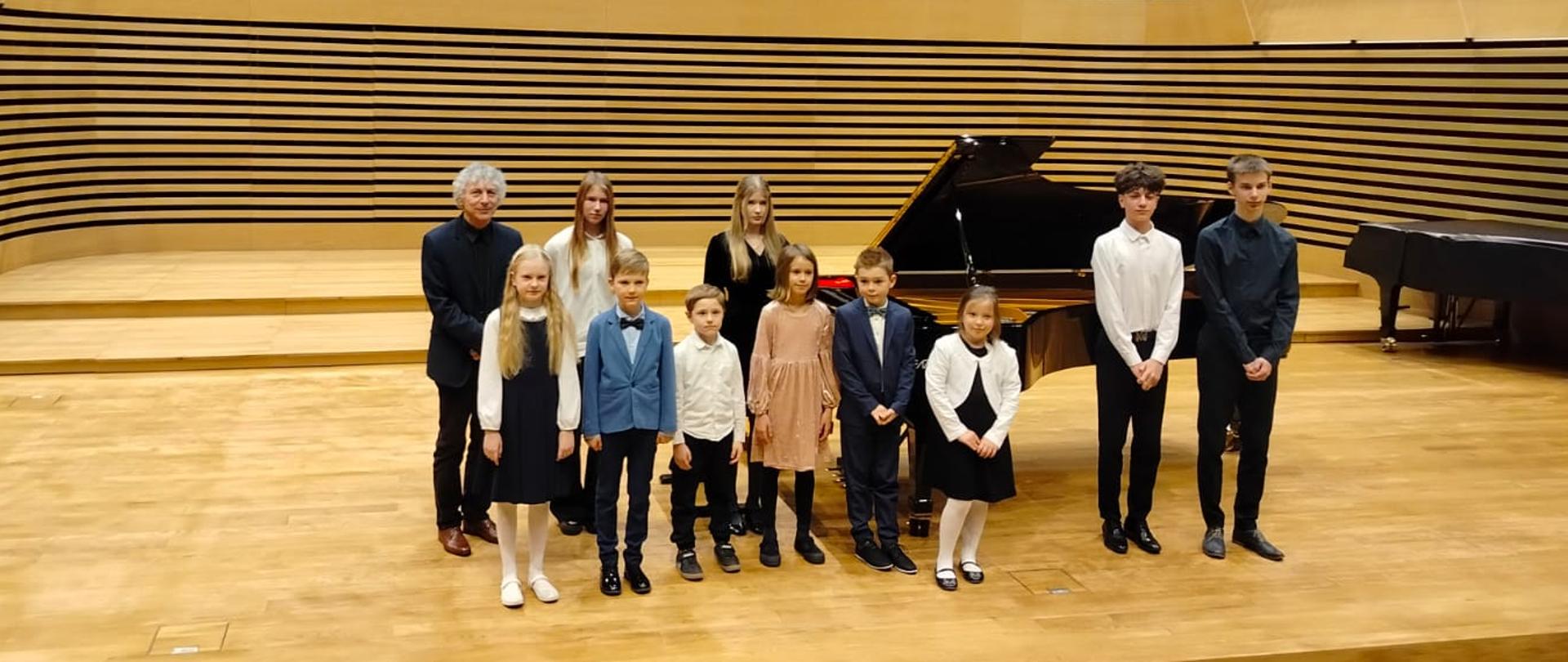 Wszyscy wykonawcy stoją z nauczycielem na estradzie, przed fortepianem.