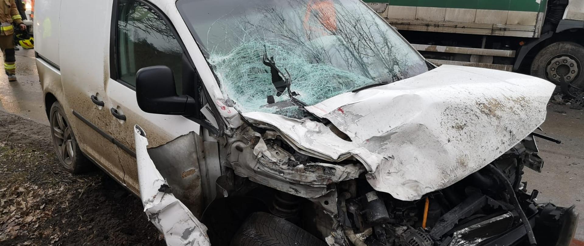 Fotografia przedstawia ujęcie wykonane z pobocza drogi wzdłuż w kierunku pasa. Na pierwszym planie samochód osobowy „dostawczak” z rozbitym przodem, oraz rozbitą przednią szybą, prawe koło pojazdu mocno uszkodzone – pęknięta felga, rozdarty błotnik. Po pojazdem widoczny rozsypany sorbent. Dalej za pojazdem widać samochód ciężarowy oraz sylwetki strażaków. Jest dzień, pochmurno.