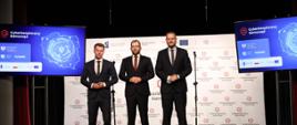 Minister funduszy i polityki regionalnej Grzegorz Puda, po jego prawej stronie stoi minister cyfryzacji Janusz Cieszyński, po lewej prezydent Otwocka Jarosław Margielski, za ich plecami baner z multiplikacją "Cyberbezpieczny Samorząd", a po lewej i prawej stronie ekrany z mapą Polski i napisem "Cyberbezpieczny Samorząd" na konferencji inaugurującej projekt „Cyberbezpieczny Samorząd”