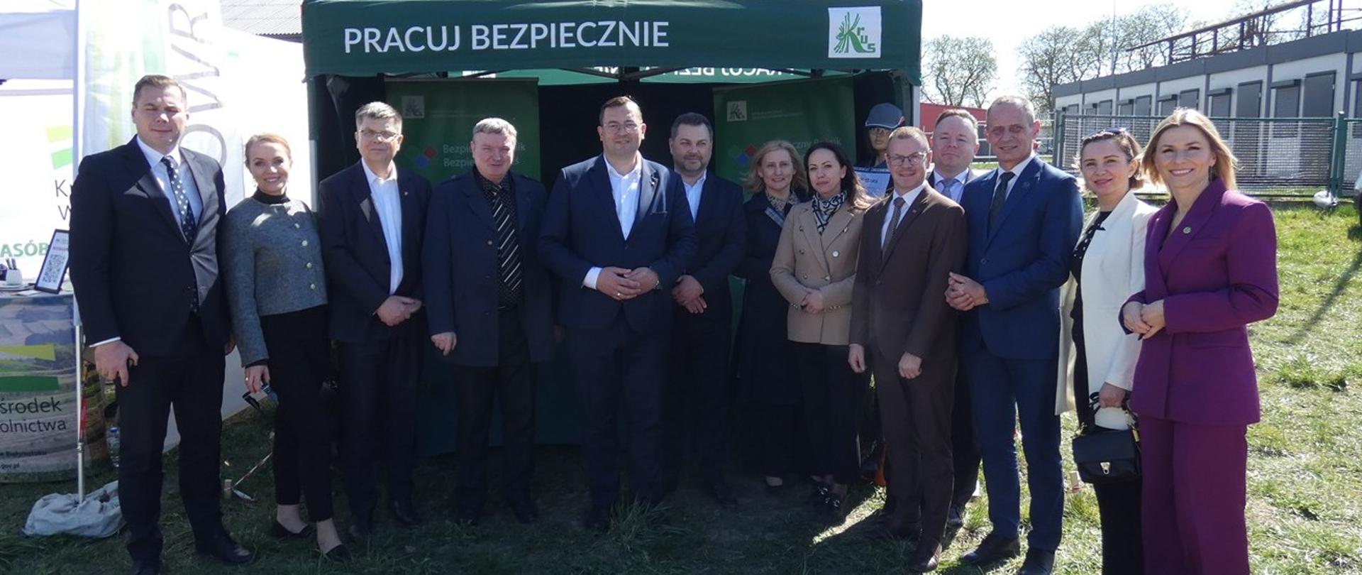 Grupa kilkunastu osób w strojach wizytowych pozuje do zdjęcia na trawie przed zielonym namiotem wystawowym z białym napisem „Pracuj bezpiecznie” i logo KRUS. 