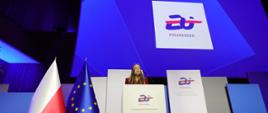 Minister funduszy i polityki regionalnej Katarzyna Pełczyńska-Nałęcz stoi na scenie za białą mównicą z napisem "EU Poland25.eu", obok niej po lewej stronie flagi Polski i UE, za jej plecami duży niebieski ekran z napisem "EU Poland25.eu"