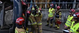 Strażacy Państwowej Straży Pożarnej w piaskowych mundurach bojowych i czerwonych hełmach prowadzą akcję ratowniczą przy uszkodzonym, pionowo postawionym samochodzie. Grupa ratowników współpracuje przy ewakuacji poszkodowanego lub rozcinaniu karoserii. Nad nimi, za metalową barierką ochronną, zmagania uważnie obserwuje publiczność, w tym inni strażacy w ubraniach wyjściowych/koszarowych oraz dzieci. W tle widoczny jest nowoczesny budynek z przeszkleniami.