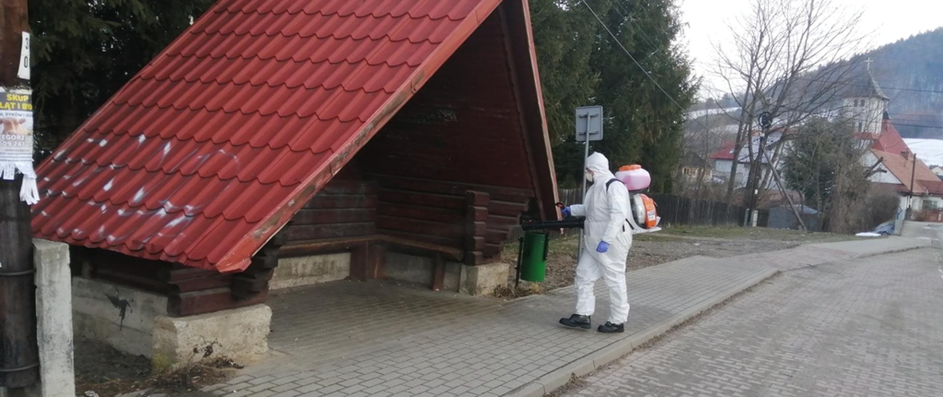 Na zdjęciu widoczny strażak w ubraniu ochronnym koloru białego w maseczce na plecach opryskiwacz spalinowy dezynfekuje wiatę przystankową środkiem odkażającym. Wiata drewniana pokryta blachą koloru brązowego.