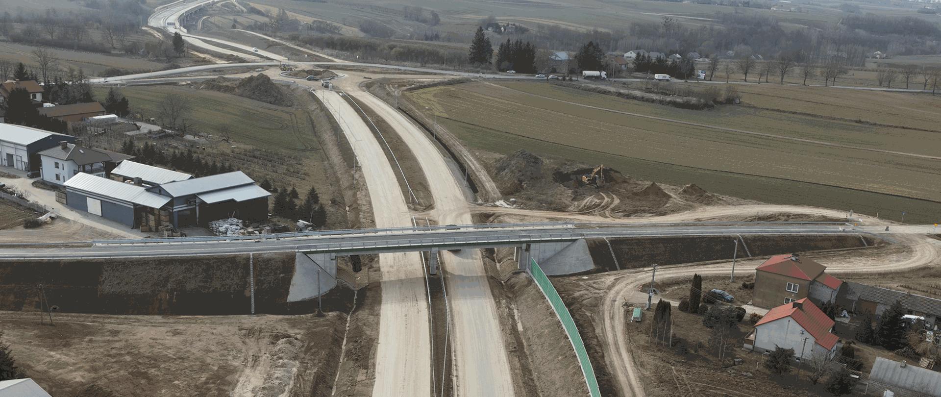 Budowa obwodnicy Opatowa - wiadukt w Tomaszowie - zdjęcie z drona - jednojezdniowy wiadukt nad dwujezdniową drogą, wokoło zabudowania, w oddali budowane rondo 