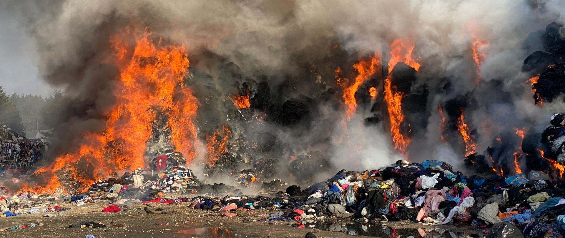 Paląca się pryzma materiałów tekstylnych. Widoczne porozrzucane materiały tekstylne. W tle widoczna kolejna pryzma materiałów tekstylnych sprasowanych w belach. Pryzma objęta ogniem i unoszący się gęsty dym. 