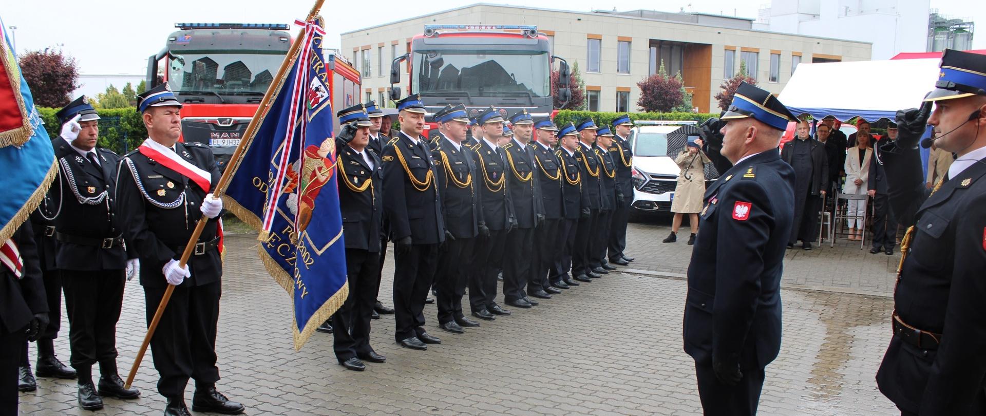 Na pierwszym planie, po lewej stronie, widoczny jest poczet sztandarowy . Obok niego w dwóch rzędach, stoi grupa strażaków w mundurach galowych, ustawionych w szyku. Po prawej stronie, naprzeciw nich, stoi strażak oddający honory. W tle widać dwa czerwone wozy strażackie oraz grupę gości, częściowo stojących pod namiotem.