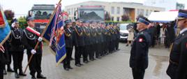 Na pierwszym planie, po lewej stronie, widoczny jest poczet sztandarowy . Obok niego w dwóch rzędach, stoi grupa strażaków w mundurach galowych, ustawionych w szyku. Po prawej stronie, naprzeciw nich, stoi strażak oddający honory. W tle widać dwa czerwone wozy strażackie oraz grupę gości, częściowo stojących pod namiotem.