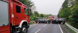 Fotografia przedstawia ujęcie wzdłuż drogi wojewódzkiej numer 156. Po lewej stronie kadru tyłem i prawym bokiem stoi samochód gaśniczy. Dalej na pasie jezdni widoczne wraki zderzonych pojazdów stojące naprzeciwko siebie w poprzek drogi, pojazdy mają rozbite przody. Za wrakami pojazdów swoje czynności wykonują strażacy w ubraniach specjalnych i w hełmach. Za uszkodzonymi pojazdami i strażakami widoczny samochód ratownictwa technicznego. Po obu stronach jezdni stoją bariery energochłonne. W tle na obu poboczach widoczny las. Jest dzień.