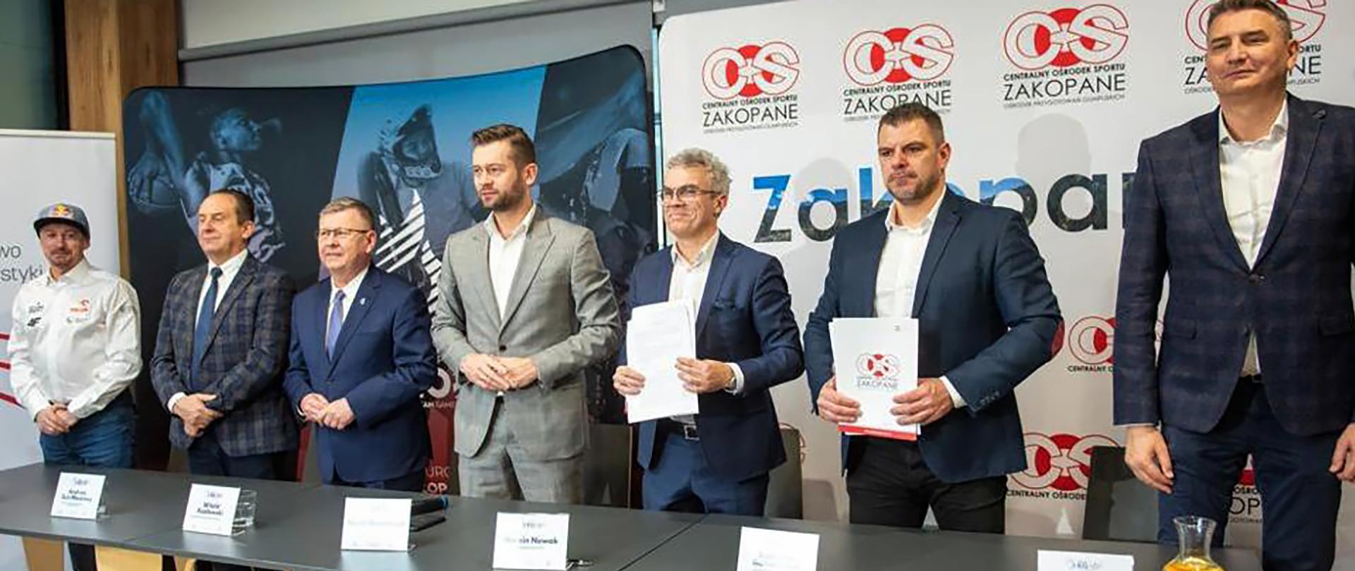 Podpisanie umowy pomiędzy COS OPO Zakopane a spółką Igrzyska Europejskie; na zdjęciu uczestnicy uroczystego podpisania umowy stoją za stołem, prezentując podpisane dokumenty