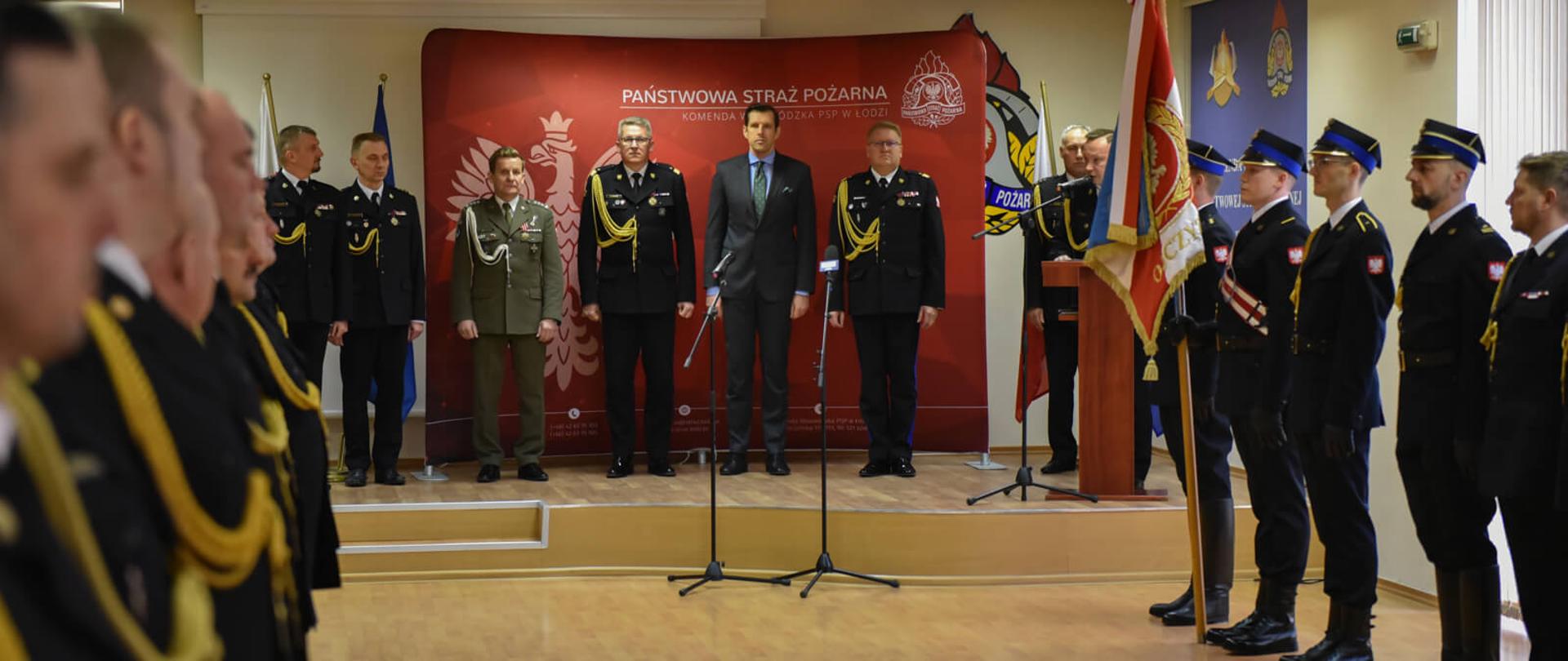 strażacy w czasie uroczystej zbiorki w sali konferencyjnej, na podwyższeniu stoją komendanci, wojewoda oraz 1 żołnierz po prawej poczet sztandarowy po lewej strażacy stojący w szeregu