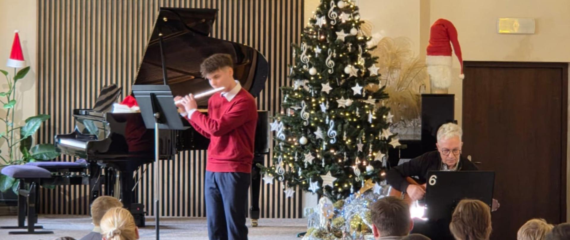 Aleksy Gurbada gra na flecie, akompaniuje mu na gitarze pan Wojciech Warszawski. Na widowni uczniowie OHP z Wolsztyna. W te dekoracje świąteczne.