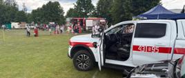 Na pierwszym planie biały, osobowy samochód straży pożarnej a drugim planie dzieci skupione wokół dużego, czerwonego pojazdu pożarniczego.