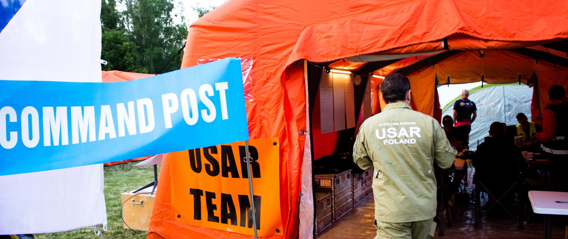 Strażak w umundurowaniu z napisem USAR Poland wchodzi do namiotu koloru pomarańczowego