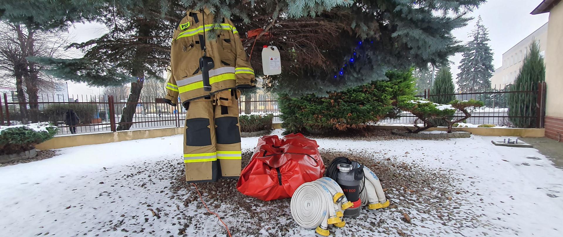 Zdjęcie przedstawia teren kamienisty pod drzewem, na którym ułożony jest sprzęt: zwinięte w krąg węże, pompa, duża czerwona paczka. Na dolnych gałęziach drzewa wisi plastikowa bańka, kamera, piaskowe spodnie i kurtka ubrania specjalnego strażackiego. W tle inne krzewy i ogrodzenie.