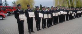 Zdjęcie przedstawia strażaków stojących na placu komendy, którzy w rękach trzymają promesy na zakup pojazdów