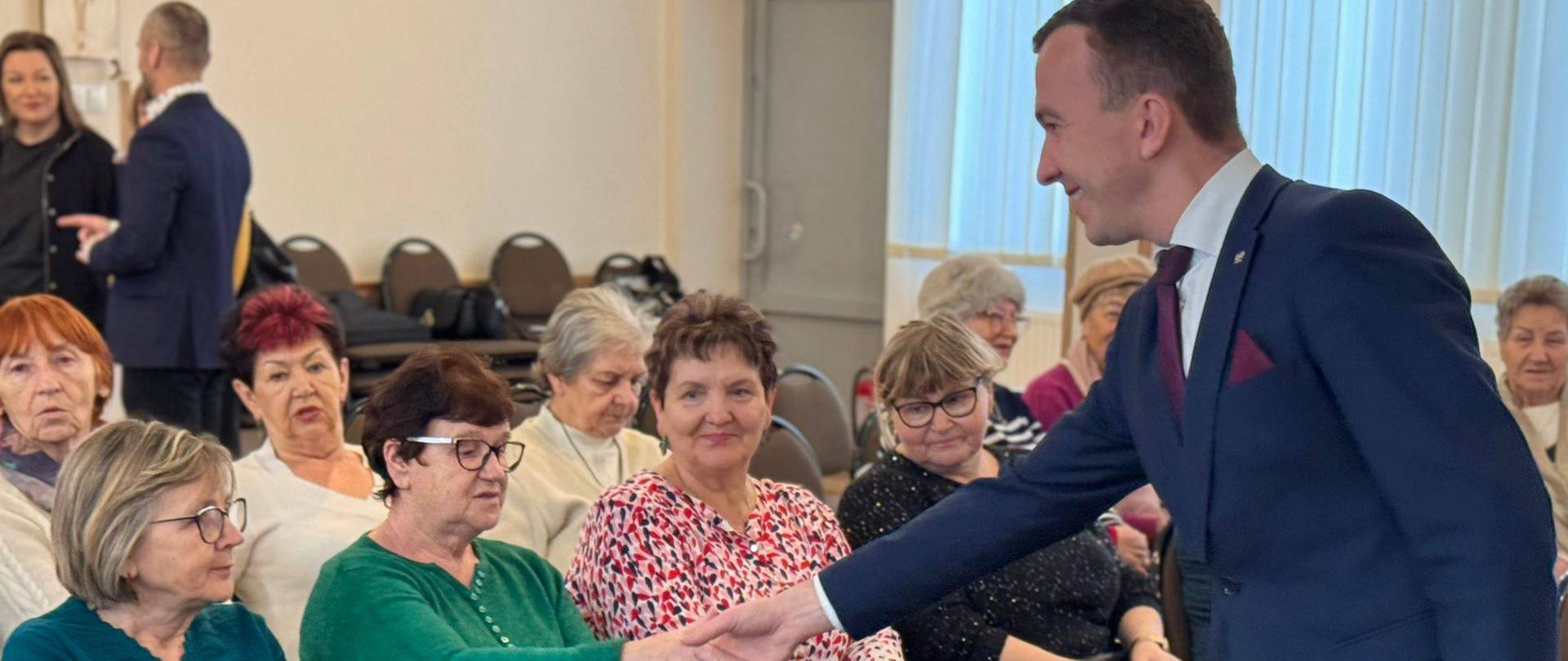 Mężczyzna w garniturze podaje rękę starszej osobie która siedzi z innymi seniorkami. Wszyscy się uśmiechają 