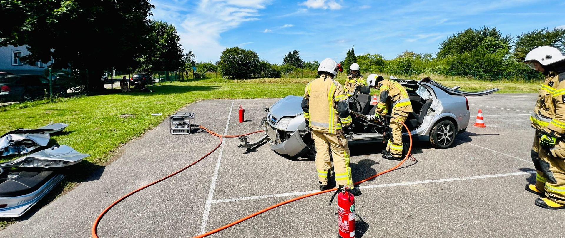Stanowisko zestawu hydraulicznego. Strażacy wykonują ćwiczenie z uwalnianiem osoby poszkodowanej zakleszczonej w samochodzie osobowym. Strażacy ubrani są w strój bojowy. Na pierwszym planie znajduje się gaśnica oraz pomarańczowe przewody z zestawu hydraulicznego