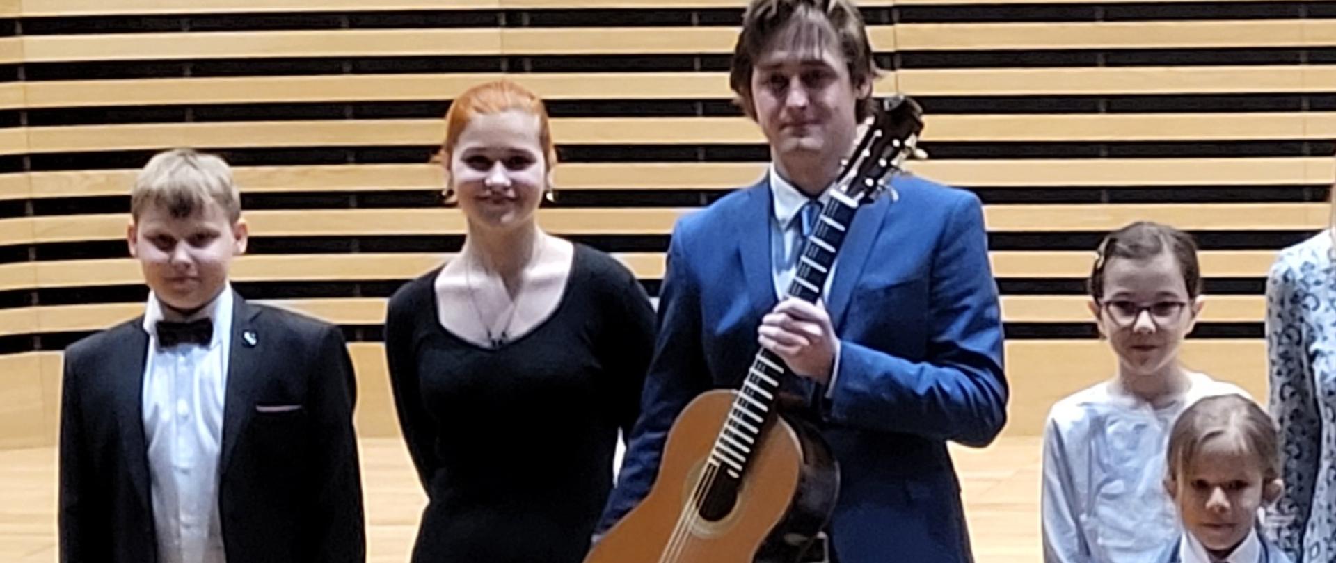 Piątka uczniów, w tym dwoje młodszych dzieci stoją w rzędzie na scenie sali koncertowej PSM, w środku stoi najstarszy uczeń, który trzyma gitarę.