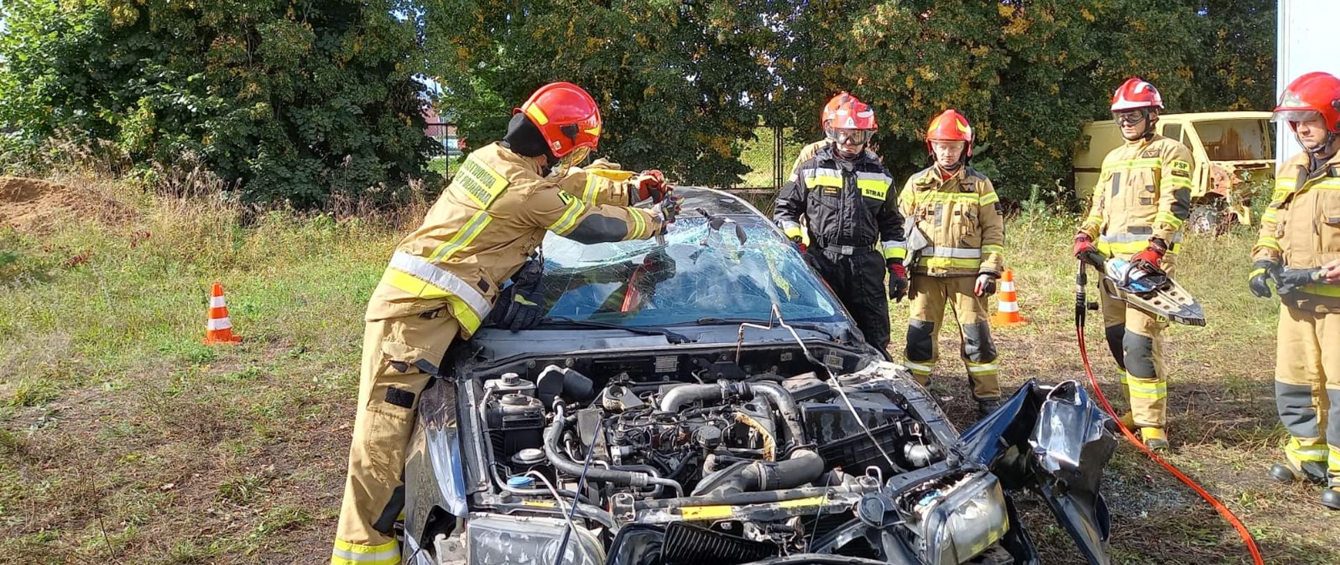 Strażak w ubraniu specjalnym rozpija przednią szybę inni strażacy stoją obok jeden ze strażaków trzyma sprzęt specjalistyczny.
