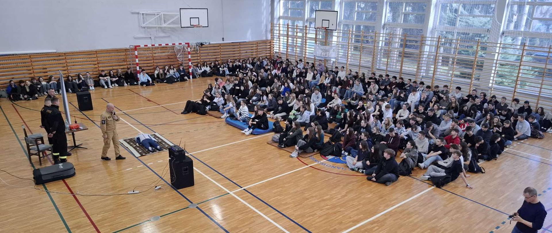 Zdjęcie przedstawia dużą salę gimnastyczną, w której odbywa się szkolenie z pierwszej pomocy prowadzone przez strażaków dla licznej grupy młodzieży. Na pierwszym planie po lewej stronie, na parkiecie, widać dwóch strażaków, z których jeden stoi przy mikrofonie, a drugi klęczy obok manekina do resuscytacji. Obok manekina leży inny manekin, prawdopodobnie dziecko. Na dalszym planie, po prawej stronie, siedzi na podłodze i na ławkach duża grupa uczniów, obserwując pokaz. W tle widoczne są duże okna, a także kosze do koszykówki i tablica wyników.