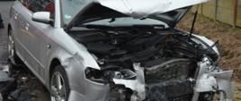 Na poboczu drogi stoi rozbity pojazd osobowy koloru srebrnego. Ma podnesioną maskę i uszkodzony przód. Z