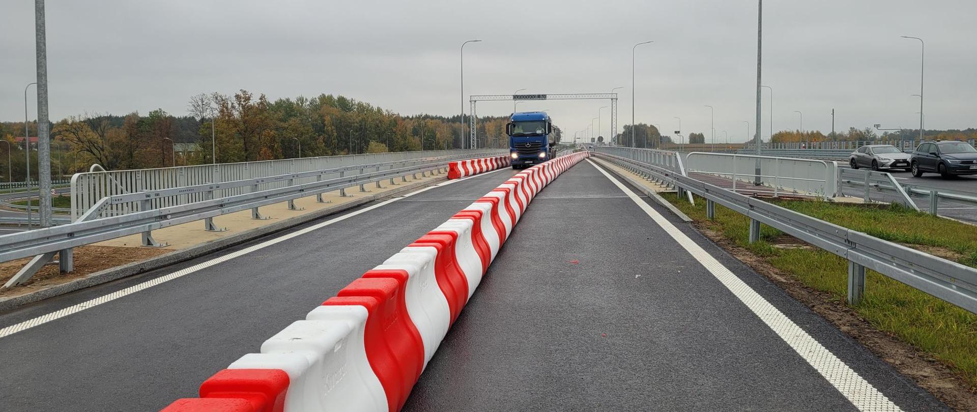 Nowo wybudowany odcinek drogi lub mostu z ciemną nawierzchnią asfaltową. Przez środek jezdni biegnie długi ciąg czerwono-białych barier separacyjnych. W oddali widoczna jest niebieska ciężarówka jadąca w kierunku aparatu. Po obu stronach drogi znajdują się metalowe bariery ochronne i wysokie latarnie. W tle widoczne są drzewa o jesiennych barwach pod pochmurnym, szarym niebem.
