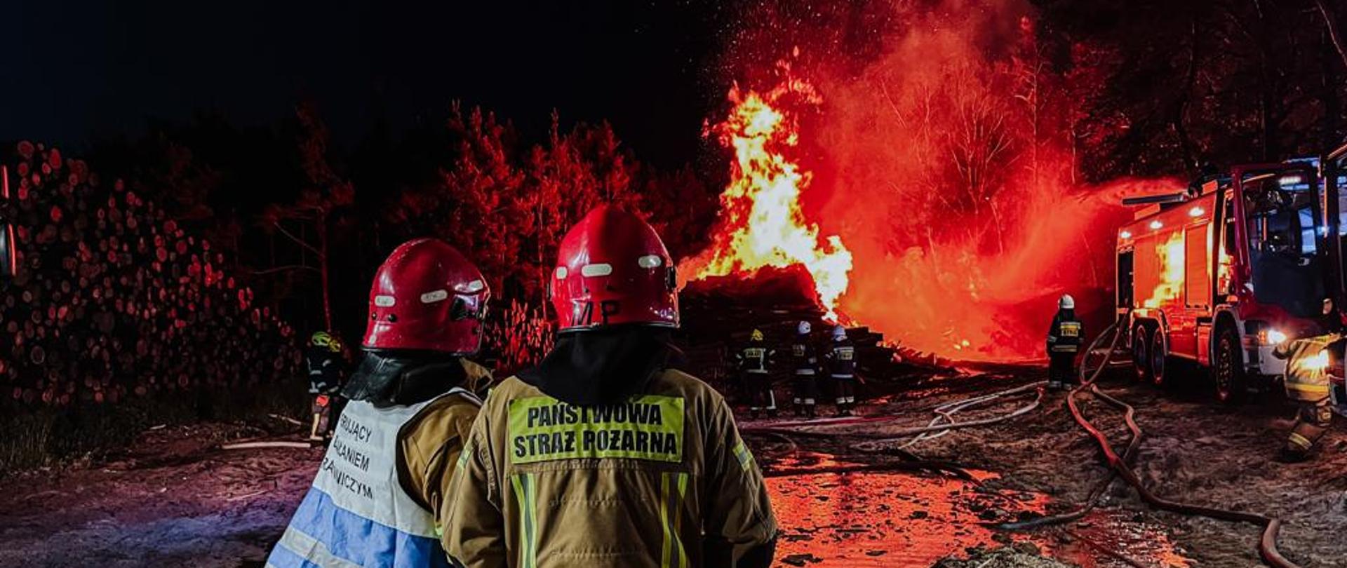 Na pierwszym planie strażacy w ubraniach specjalnych kontrolują działania gaśnicze i rozwój pożaru. Na dalszym planie widać rozwinięte węże pożarnicze od samochodu. W tle widać palący się skład drewna i strażaków, którzy gaszą pożar.