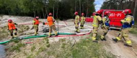 Strażacy przy zbiorniku wodnym w lesie podczas ćwiczeń. W tle samochód pożarniczy oraz przyczepka wężowa.