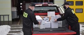 Strażacy wypakowują z pojazdu strażackiego typu pick-up środki ochrony indywidualnej do walki z Covid-19 (m.in. maski ochronne i płyny do dezynfekcji). Strażacy są zabezpieczeni w maski ochronne. 
