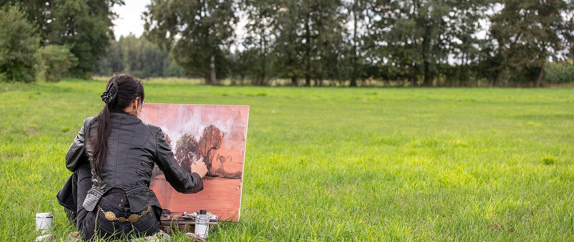 Na zdjęciu widać osobę, która siedzi na trawie i maluje na dużym płótnie. Ma na sobie czarną, skórzaną kurtkę. Jest odwrócona plecami, więc widać, jak jej ręka z pędzlem porusza się po płótnie. Obok niej leżą słoiki z farbami. Scena jest na dużej, otwartej łące z drzewami w tle.