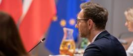 Wiceminister Waldemar Buda siedzi przy stole konferencyjnym, przed nim flagi Polski i Czech