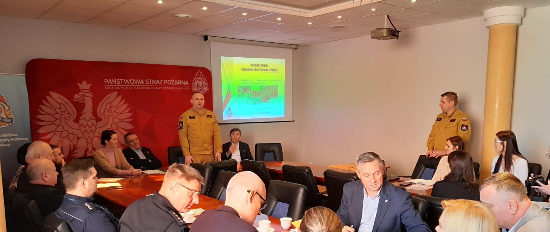 Zdjęcie w pomieszczeniu. Oświetlenie naturalne. Widać członków komisji i zespołu zarządzania kryzysowego oraz funkcjonariuszy PSP. Meble biurowe, drewniane, brązowe.