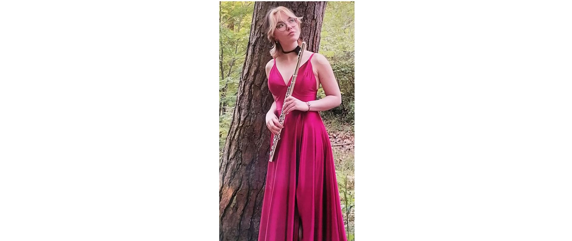 Zdjęcie kolorowe. Dziewczyna w czerwonej sukni, trzyma flet. W tle pień drzewa.