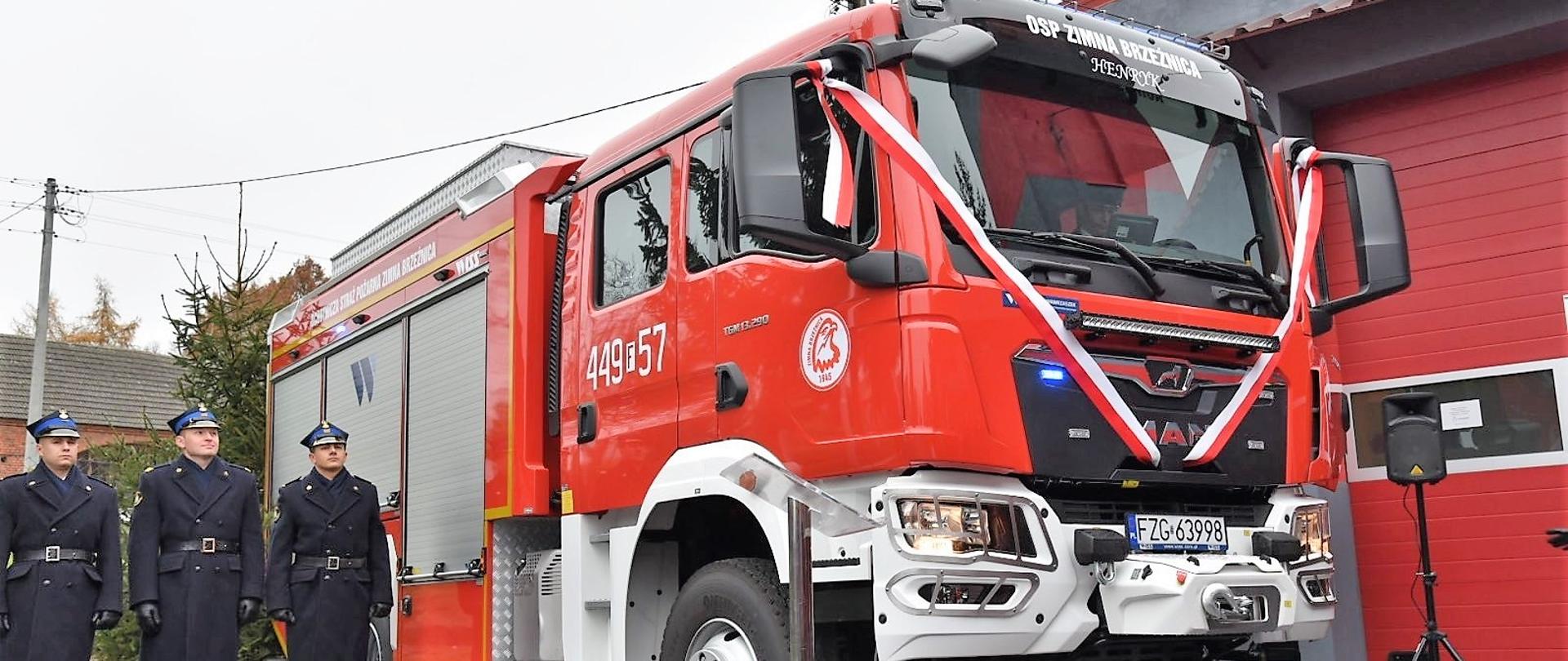 Obok nowego wozu przewiązanego biało-czerwoną szarfą stoi 3 druhów w galowych mundurach 