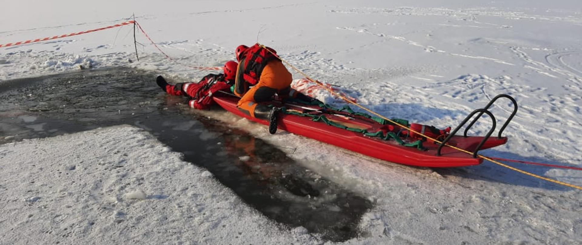 Strażak w skafandrze wypornościowym podejmuje przy użyciu sań lodowych osobę poszkodowaną z przerębla na terenie akwenu zalodzonego